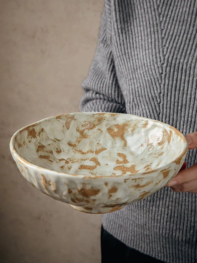 Rustic stoneware serving bowl with hand-painted amber brushstroke glaze, held against neutral beige background, interior view showing cream and amber reactive glaze pattern