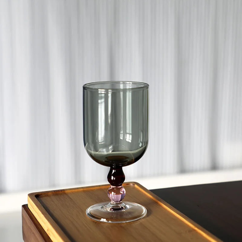 "Chromatic Orb" glass goblet in smoke grey with two-tone wine and pink bead stem, on a wooden tray against a white curtain background