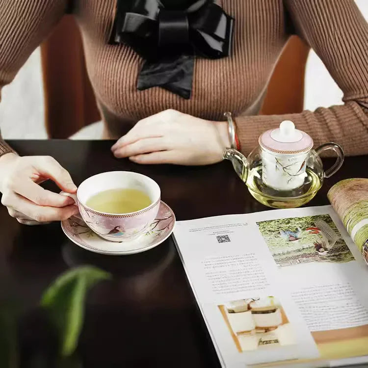 Woman in a brown ribbed jumper holding a pink "Happy Bird" porcelain tea cup and saucer at a dark table, with the glass teapot and an open magazine in the foreground.
