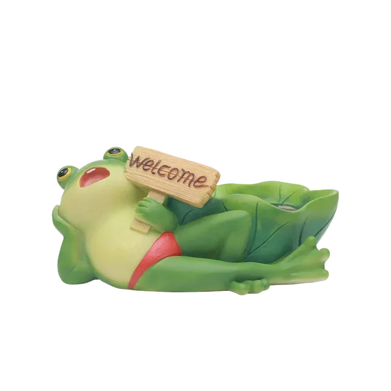 "Frog Tale" resin frog planter in Welcome Lounger — hand-painted frog reclining on a lily leaf holding a wooden Welcome sign, white background
