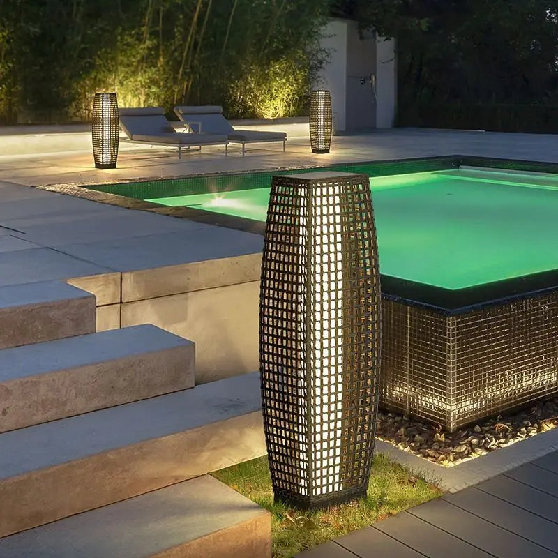 Dark wicker solar floor lanterns glowing beside a swimming pool at night, placed along a stone terrace with sun loungers and bamboo garden in the background