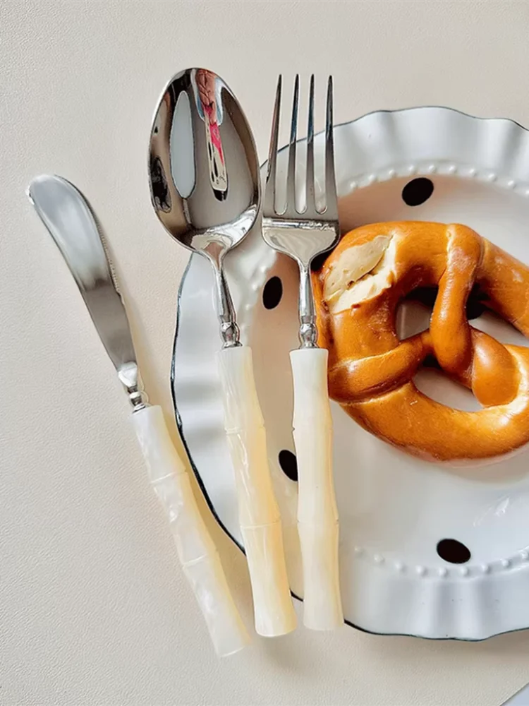 "Jade Stem" Ivory Cream cutlery set — knife, dinner spoon and fork styled beside a ceramic polka dot plate with a pretzel — casual lifestyle shot showing everyday use