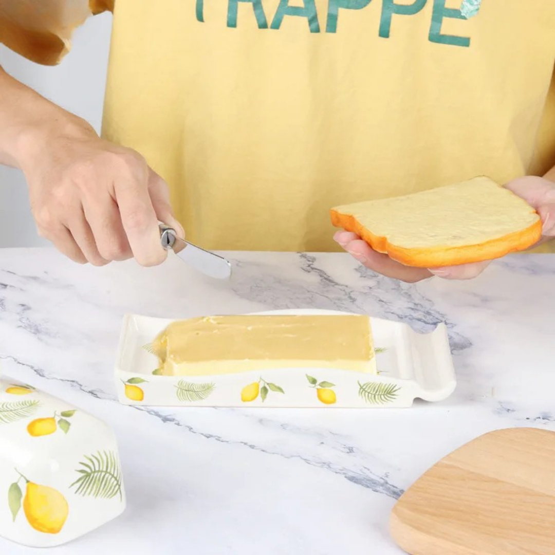 Hands spreading butter on a slice of bread using a knife, with a lemon-patterned ceramic butter dish tray on a marble surface and the domed lid set aside.