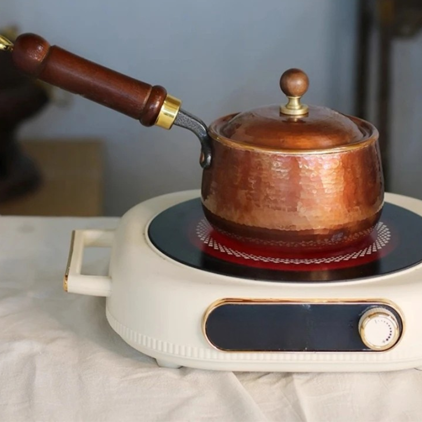 Copper Forge saucepan with lid closed heating on a vintage cream and gold induction hob with glowing red element visible beneath — hammered copper body and wooden knob lid detail, white linen cloth background with soft blue-grey wall — induction compatibility lifestyle shot by CL House