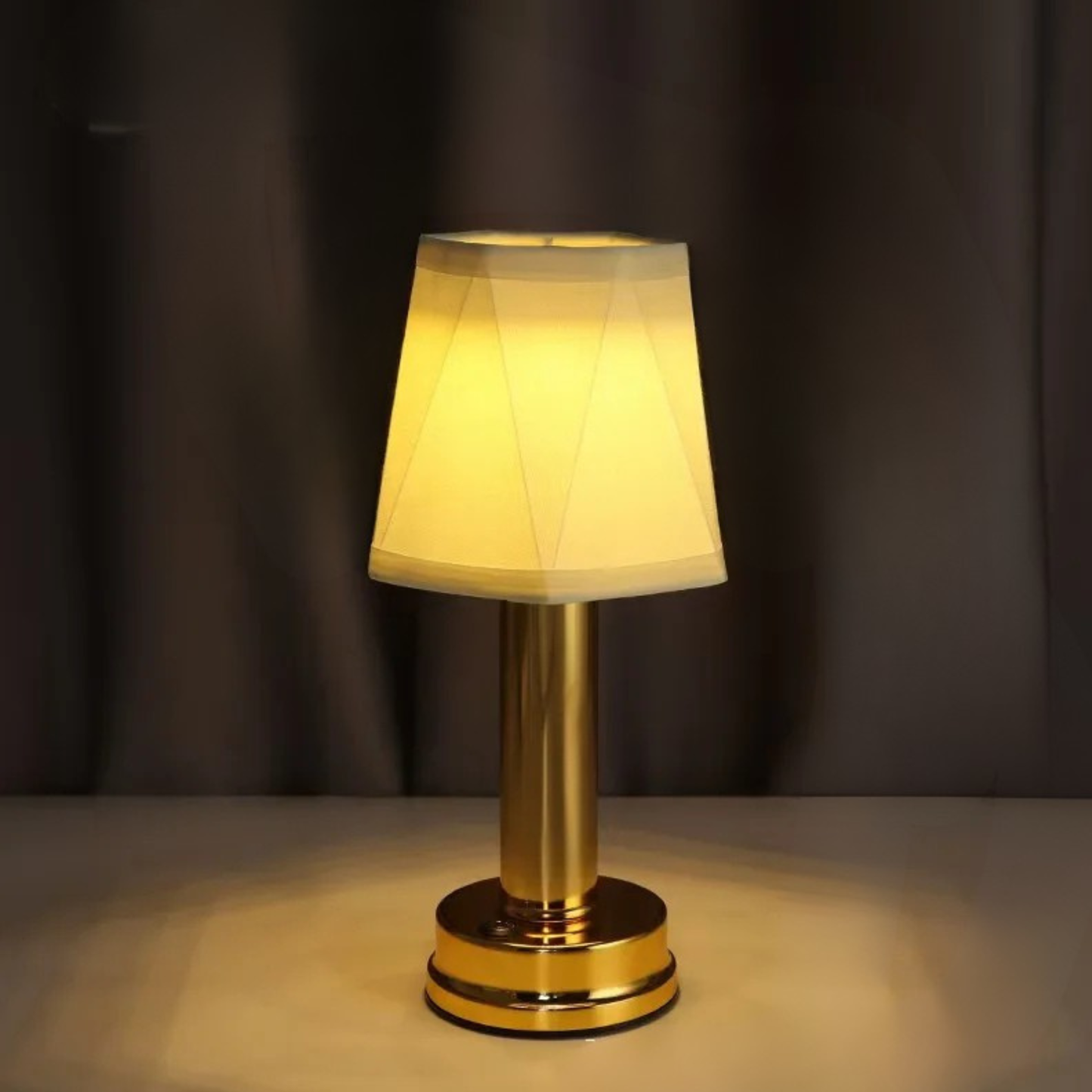 Gold rechargeable touch table lamp with pleated fabric shade glowing warm amber light against dark curtain backdrop, creating a cosy intimate atmosphere