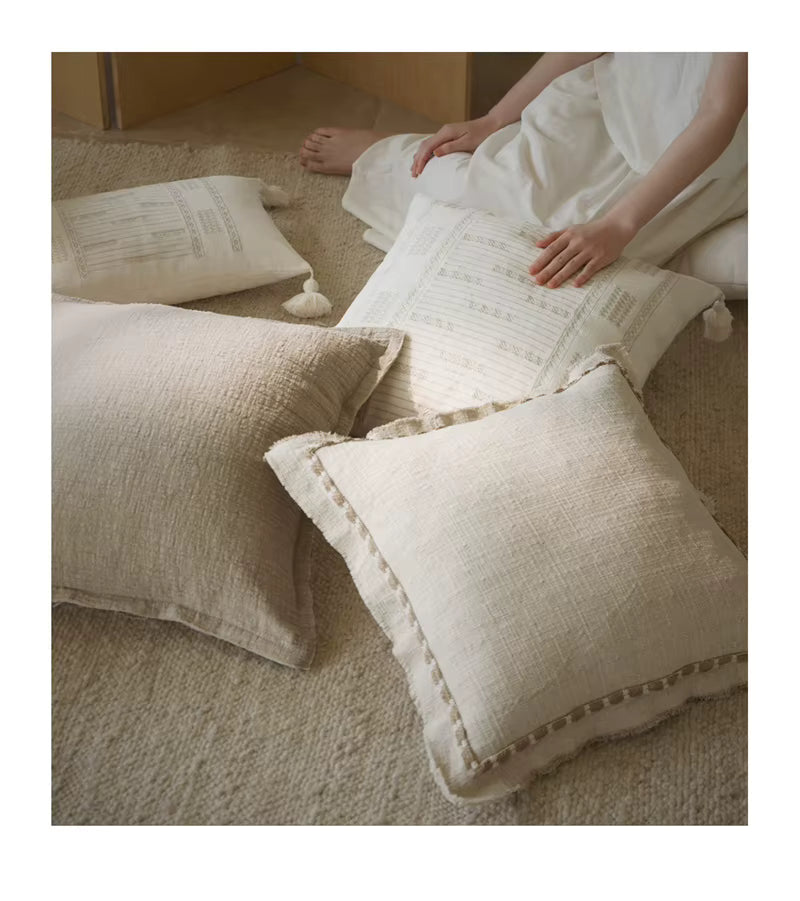 Woman in white linen outfit arranging ivory and natural linen cushion covers with tassels and fringe edges on a textured jute rug, styled in a calm wabi-sabi inspired setting.