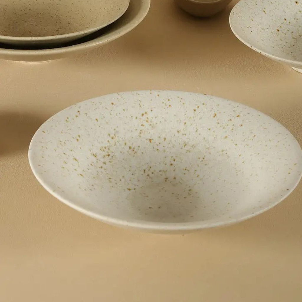 Speckled clay handcrafted ceramic dinner plate in off-white with natural earth tones, shown close up on a beige surface with additional stacked plates in the background
