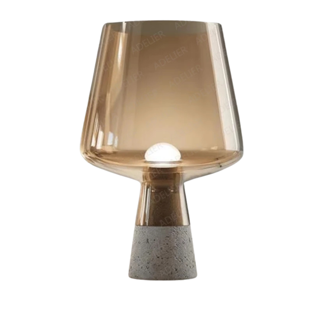 A modern table lamp with a wide amber glass shade glowing with warm light, standing on a tapered grey speckled cement base against a dark background.