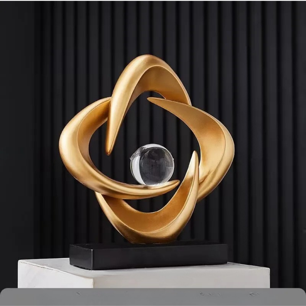 Gold abstract resin sculpture with three interlocking curved loops and a clear crystal sphere on a black rectangular base, set against a dark ribbed backdrop.