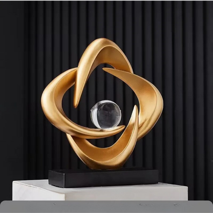 Gold abstract resin sculpture with three interlocking curved loops and a clear crystal sphere on a black rectangular base, set against a dark ribbed backdrop.