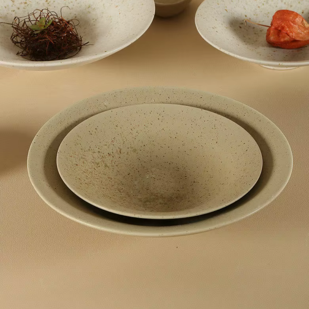 Speckled clay handcrafted ceramic dinner plates stacked in two sizes on a beige surface, with additional plates styled with dried botanicals and orange physalis in the background