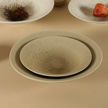 Speckled clay handcrafted ceramic dinner plates stacked in two sizes on a beige surface, with additional plates styled with dried botanicals and orange physalis in the background