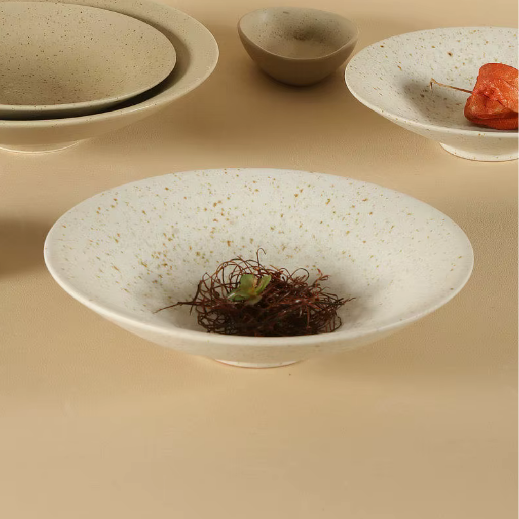 Speckled clay handcrafted ceramic pasta bowl in off-white with dried botanical nest and small succulent inside, surrounded by stacked plates, a small ceramic bowl and a plate with dried orange physalis in the background