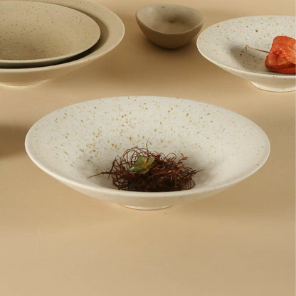 Speckled clay handcrafted ceramic pasta bowl in off-white with dried botanical nest and small succulent inside, surrounded by stacked plates, a small ceramic bowl and a plate with dried orange physalis in the background