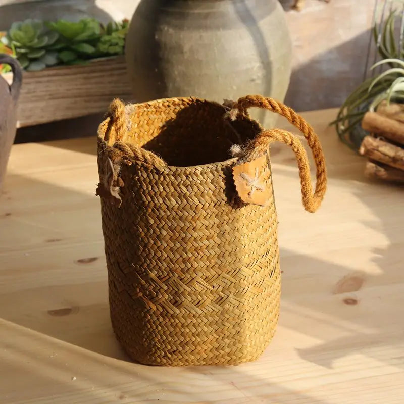 Small natural yellow woven straw flower basket vase with rope handles and decorative leaf details displayed on a wooden table.