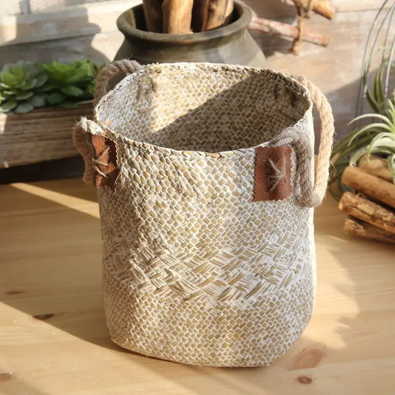

Large white woven straw flower basket vase with rope handles and decorative leaf details displayed on a wooden table.