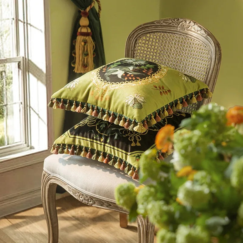  "Tropical Reverie" velvet cushion cover designs stacked on a vintage carved chair — Jungle Sovereign in sage green above, Midnight Hummingbird in dark ground below, multicolour tassel trim, natural window light
