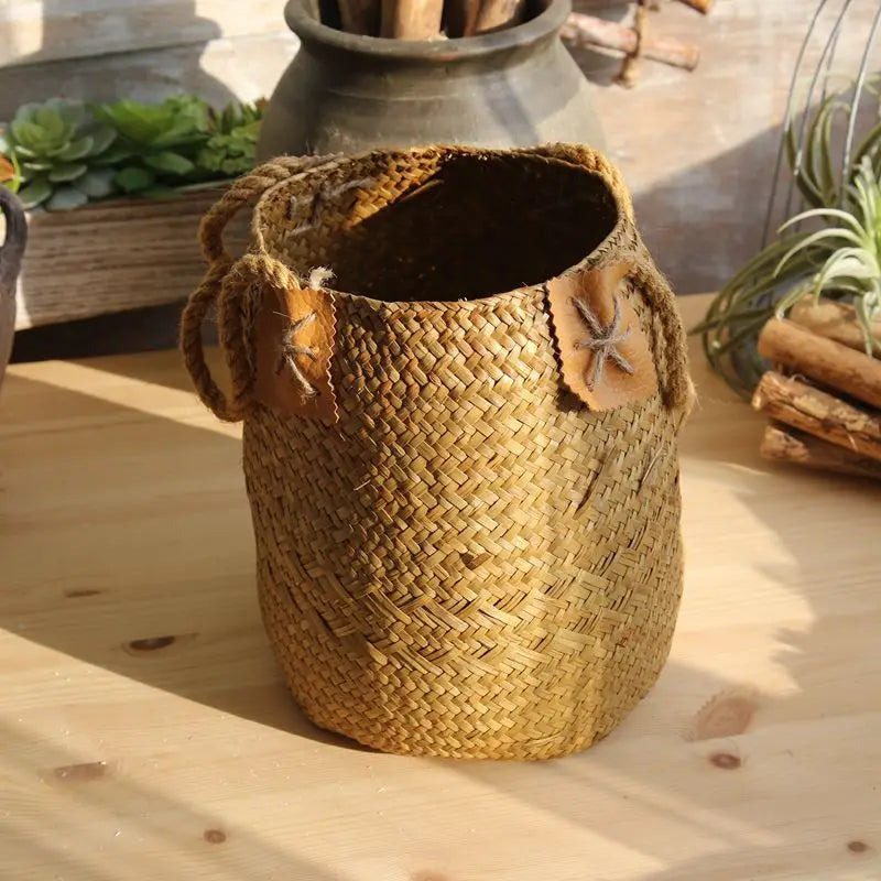 Texto alternativo: Large natural yellow woven straw flower basket vase with rope handles and decorative leaf details displayed on a wooden table.