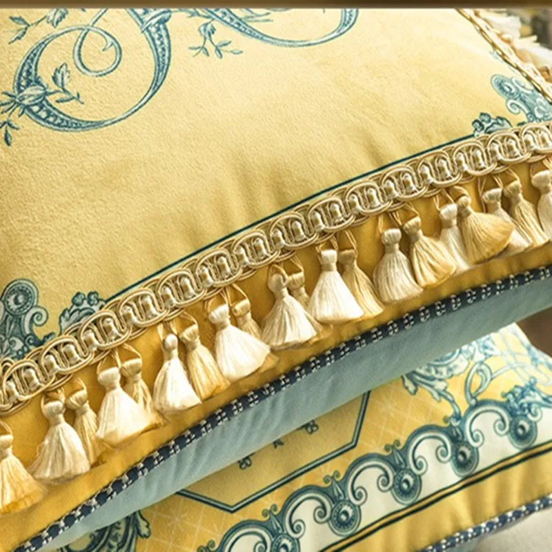: Close-up of velvet cushion cover edge — golden braided trim with cream tassel fringe, teal baroque print visible on golden yellow velvet ground