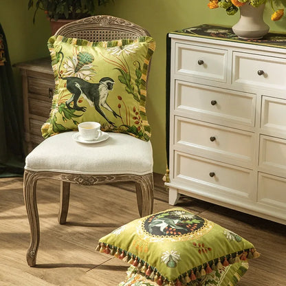 "Tropical Reverie" velvet cushion cover styled on a vintage carved chair — Jungle Sovereign design with monkey and tropical botanicals, alongside the medallion variant on the floor, sage green interior setting