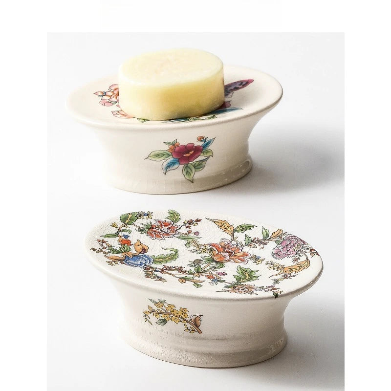 Two oval pedestal floral ceramic soap dishes stacked — Butterfly Garden variant holding a natural bar of soap on top, Chinoiserie Garden variant below, on grey background