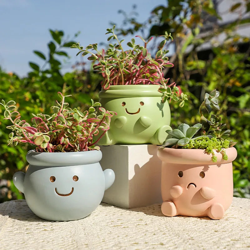 "Happy Pot" set of three smiley face resin succulent planters in sky blue, sage green and blush pink, displayed outdoors with lush succulents and green garden background
