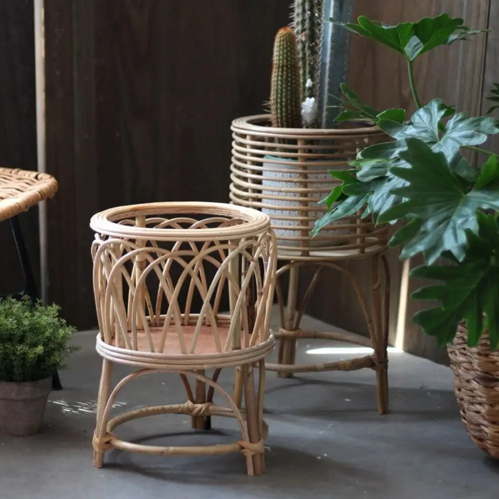 Rattan Weave" natural rattan plant stands styled in an outdoor garden setting — Arch Weave Small in foreground and Spiral Weave Large with cactus in background, surrounded by tropical plants, warm natural tone finish