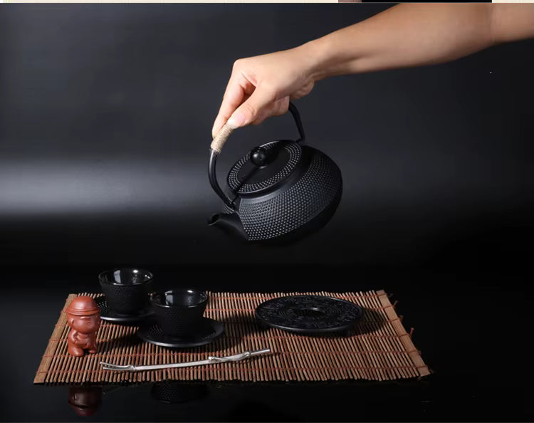  Hand lifting a cast iron teapot, Iron Black finish — hobnail texture, styled with Japanese tea ceremony set on bamboo mat, dark background — Iron Ritual by CL House