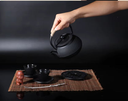  Hand lifting a cast iron teapot, Iron Black finish — hobnail texture, styled with Japanese tea ceremony set on bamboo mat, dark background — Iron Ritual by CL House