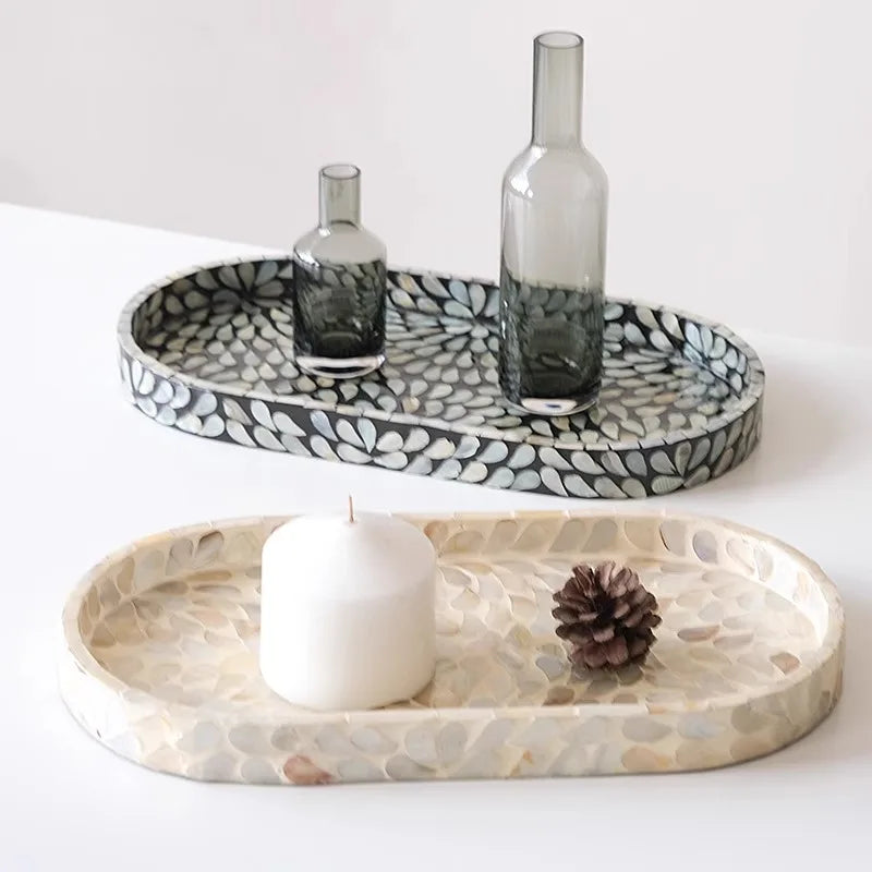Two mother-of-pearl oval trays side by side — Midnight black shell mosaic tray with smoky glass vases above, Pearl cream shell mosaic tray with white candle and pine cone below — artisan bathroom decor