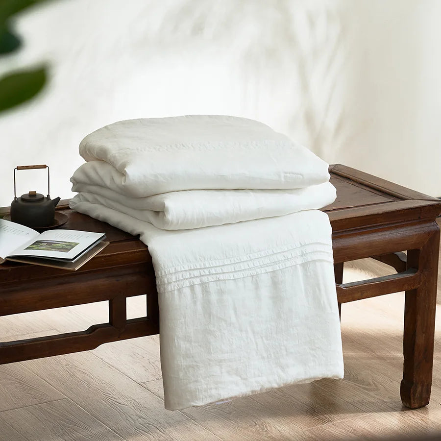  Ivory white quilted linen cotton summer throw blanket, neatly folded on a dark wood bench in a japandi-style interior with wooden shelving and open book