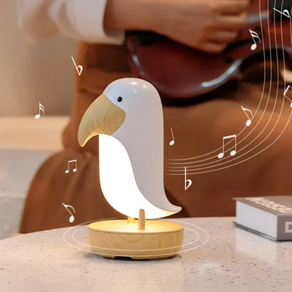 Birdie LED night light glowing on a marble surface next to a book, with music notes illustrating the Bluetooth speaker while someone plays guitar in the background