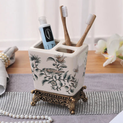  White ceramic toothbrush holder with delicate garden bird and botanical branch pattern in sage green and grey, ornate bronze metal base, two bamboo toothbrushes and toothpaste displayed, lifestyle background