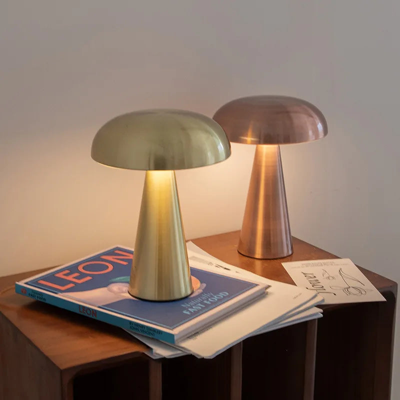 Two Cap & Glow mushroom table lamps glowing with warm amber light on a wooden side table — one in Golden and one in Rose Gold — styled alongside books and magazines in a cosy living room setting.