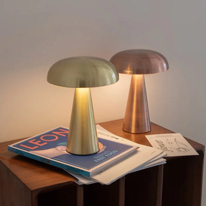 Two Cap & Glow mushroom table lamps glowing with warm amber light on a wooden side table — one in Golden and one in Rose Gold — styled alongside books and magazines in a cosy living room setting.
