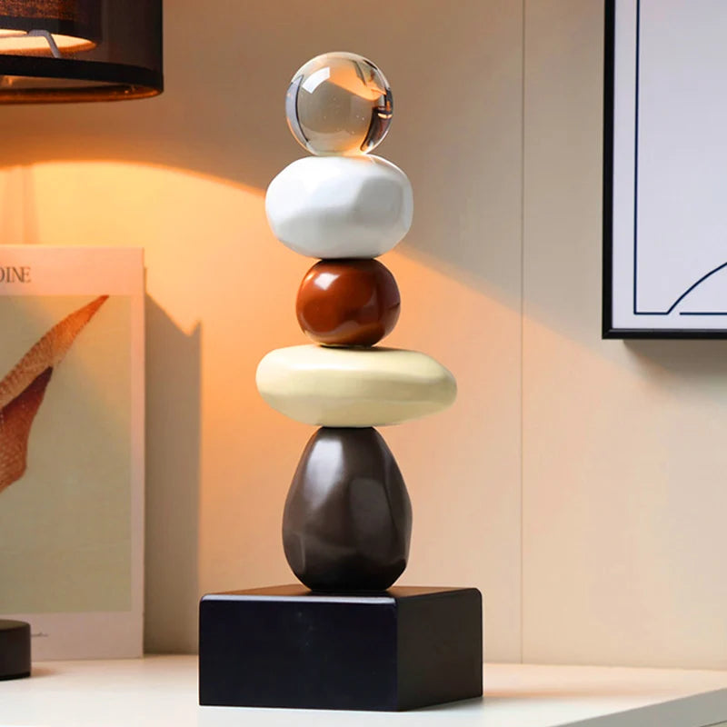 Abstract ceramic stacked sculpture with a clear upper sphere, white and brown rounded forms, a cream oval element and a black square base, displayed on a dark console in a softly lit modern interior.⠋