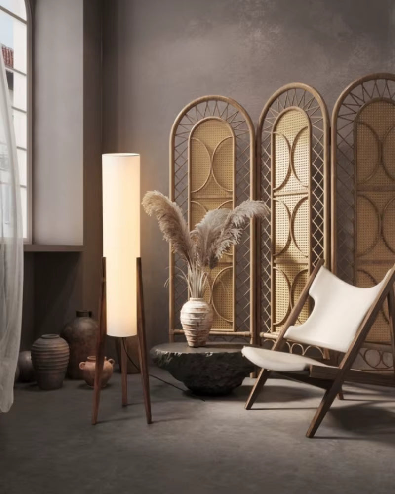 "Tripod & Cylinder" walnut floor lamp casting warm light beside a rattan room divider, pampas grass and a linen lounge chair