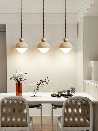 Three Stone & Orb travertine pendant lights with log wood beads clustered above a white dining table styled with rattan chairs, a red glass vase with white flowers and ceramic tableware against a neutral wall