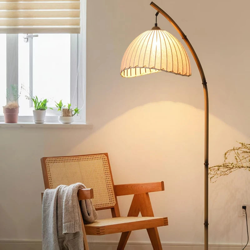 Arc & Pleat canvas floor lamp beside a rattan armchair with a knit throw, glowing warmly next to a sunlit window with small potted plants on the sill