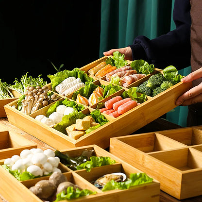 Person carrying a Bamboo Grid 9-section tray abundant with fresh hot pot ingredients — corn, seafood rolls, broccoli, sausages, tofu and mushrooms — over a table set with additional bamboo trays, green curtain backdrop