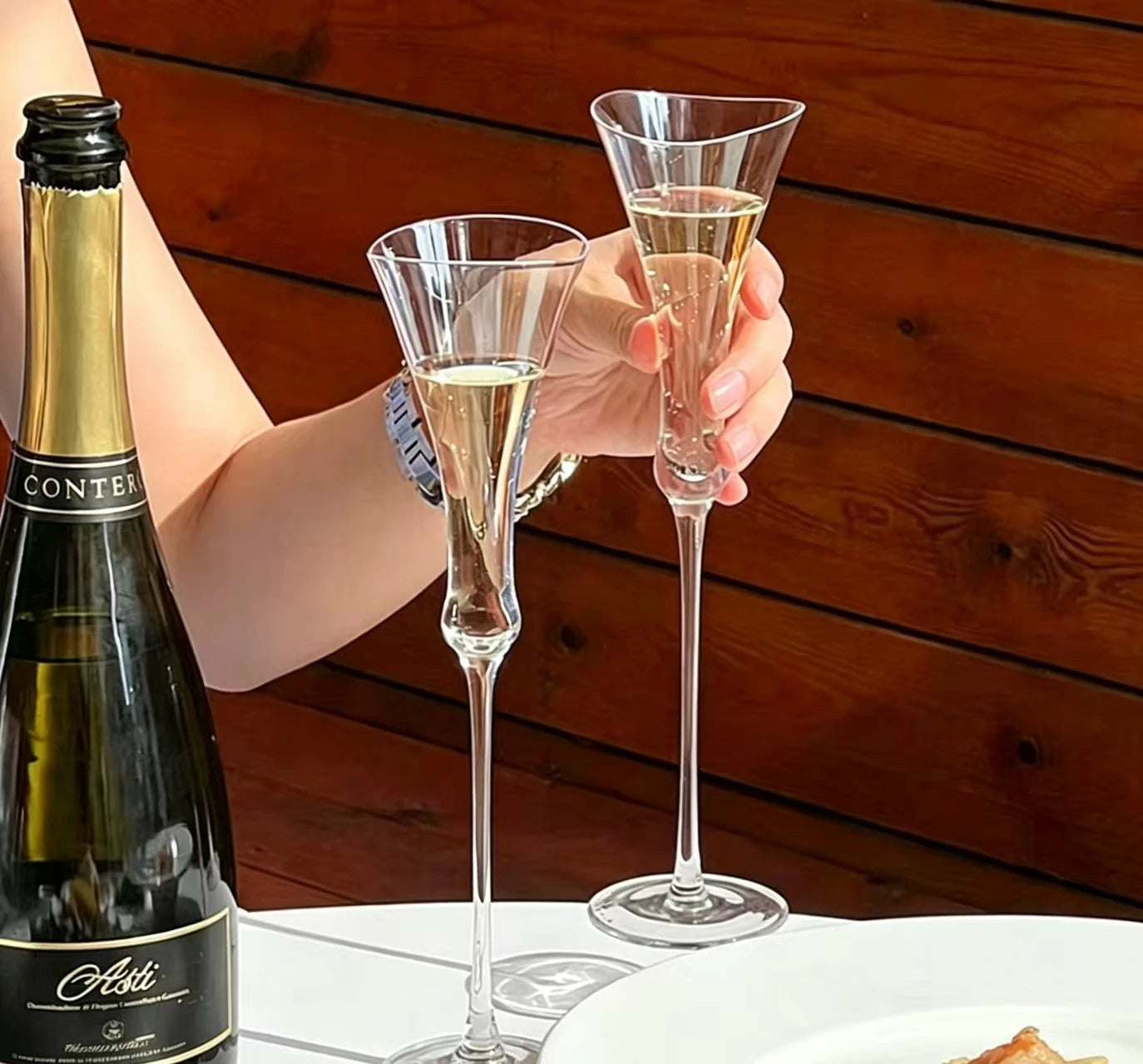 Two "Trumpet" crystal champagne flutes filled with champagne, hand holding one glass alongside a champagne bottle — CL House Drinkware collection