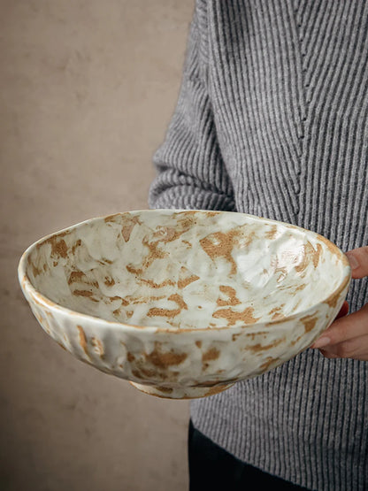Rustic stoneware serving bowl with hand-painted amber brushstroke glaze, held against neutral beige background, interior view showing cream and amber reactive glaze pattern