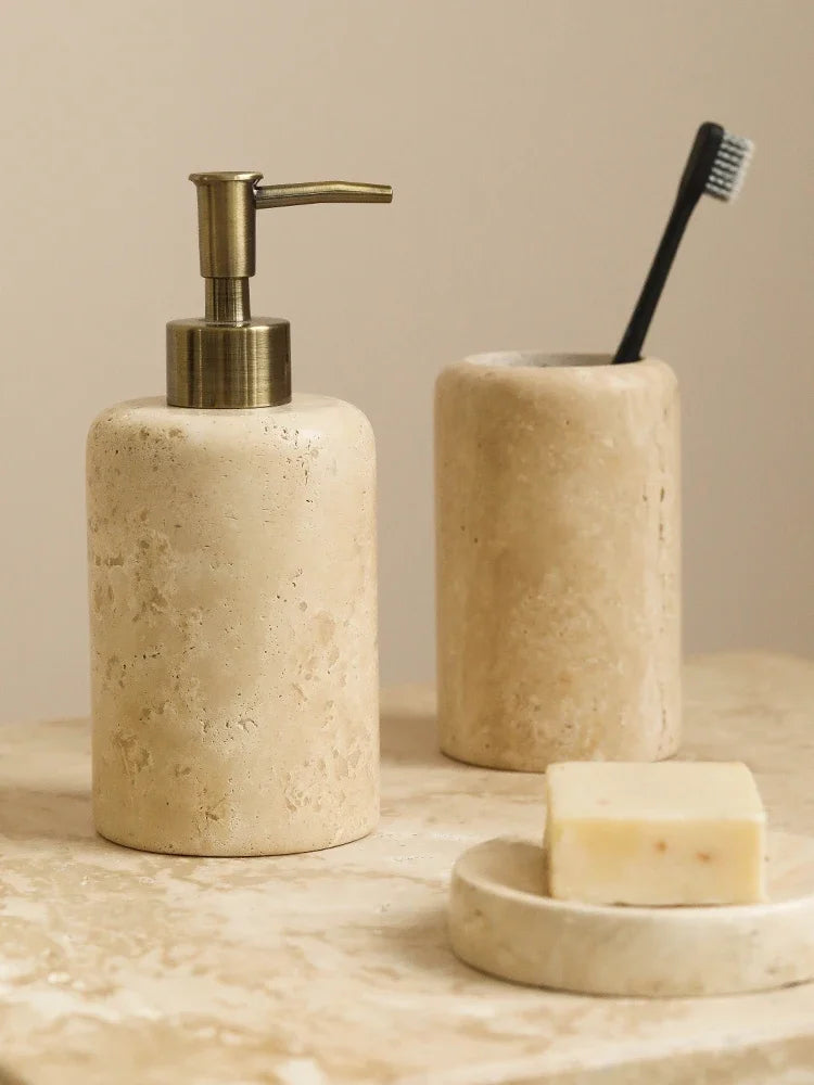  Natural travertine soap dispenser with antique brass pump alongside toothbrush cup and round soap dish, warm beige stone texture on a marble surface — japandi bathroom accessories