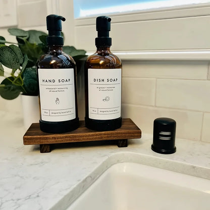  Walnut wood elevated tray beside kitchen sink holding two amber glass soap dispensers with black pumps, marble countertop and white tile background — farmhouse kitchen styling