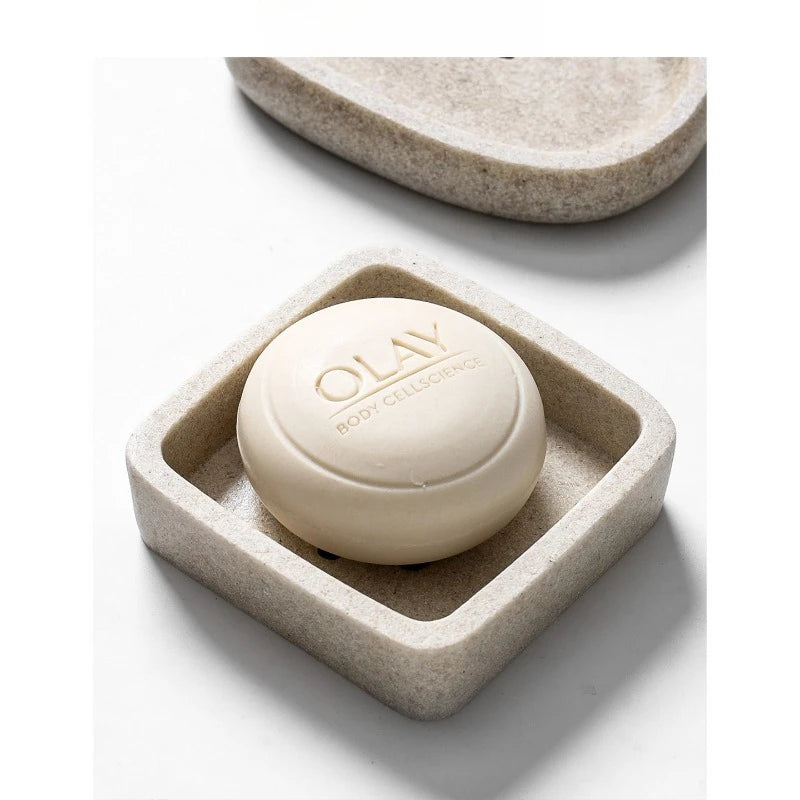 Close-up of square stone-texture resin soap dish holding a round white soap bar, warm sand beige finish with speckled detail, pebble dish visible in background