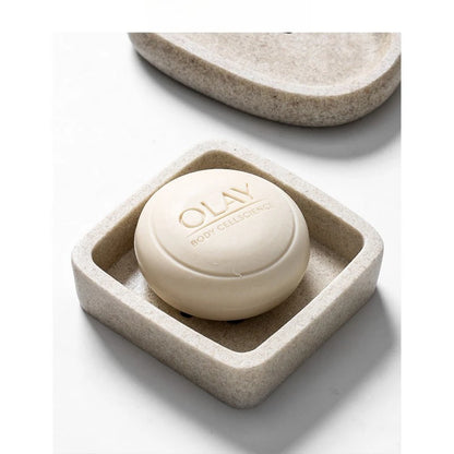 Close-up of square stone-texture resin soap dish holding a round white soap bar, warm sand beige finish with speckled detail, pebble dish visible in background