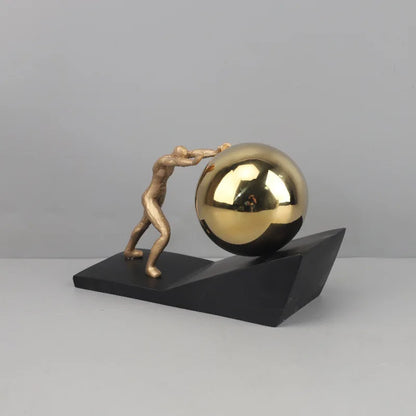 Gold Sphere version of The Ascent metal Sisyphus sculpture, with a gold figure pushing a reflective gold sphere up a black angled base.