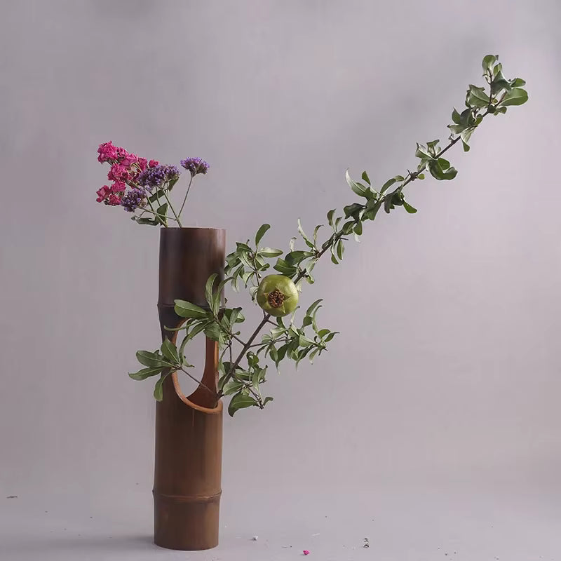 Handmade dark bamboo sculptural vase with carved opening, styled with pomegranate branch and dried pink and purple flowers against a light grey background