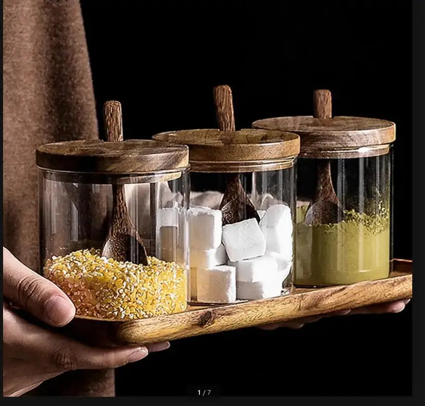 Close-up of hands holding an acacia wood tray with three glass jars filled with golden millet, white sugar cubes and green matcha powder, wooden spoons resting on each acacia lid, dark moody background — lifestyle detail of the Acacia & Glass Spice Jar Set by CL House