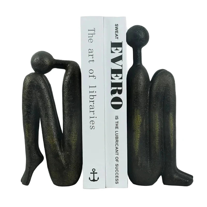 Pair of dark resin abstract human sculptures positioned on either side of two white books, with one figure leaning forward and the other standing upright.